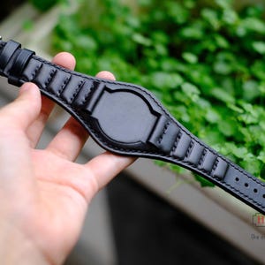 May include: Black leather watch strap with a stainless steel buckle. The strap features a padded design with decorative stitching and a central section for the watch face. The strap is held in a hand against a blurred green background.