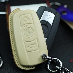 May include: A close-up of a beige leather key fob with a silver key ring and a black car key with a silver lock button. The key fob has a house icon and the word "LOCK" on it.