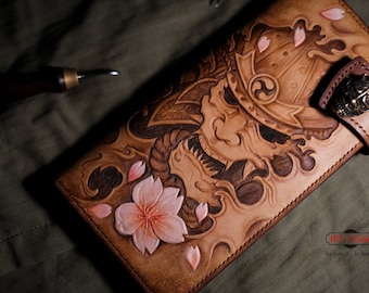 May include: A brown leather wallet with a detailed carving of a samurai mask, cherry blossoms, and waves. The wallet has a brass clasp with a smaller samurai mask design.