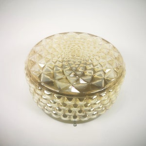 Op de afbeelding: Een ronde, amberkleurige glazen container met een getextureerd, geometrisch patroon. Het deksel en de basis hebben een herhalend ruitontwerp, wat een decoratieve en vintage esthetiek creëert. De container wordt waarschijnlijk gebruikt voor opslag of weergave.