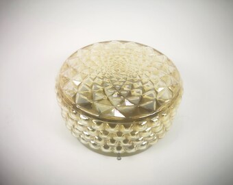 May include: A round, amber-colored glass container with a textured, geometric pattern. The lid and base feature a repeating diamond design, creating a decorative and vintage aesthetic. The container is likely used for storage or display.