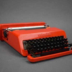 Peut inclure: Une machine à écrire vintage Olivetti Lettera 32 rouge avec des touches noires. La machine à écrire a une poignée de transport et un ruban noir.
