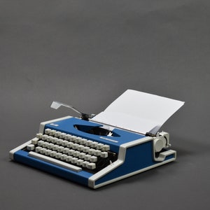 May include: A blue and white vintage typewriter with a blank sheet of paper in the carriage. The typewriter has a classic design with a round carriage return knob and a lever for adjusting the paper.