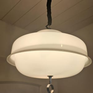 May include: A white, retro-style pendant light fixture. The light has a large, rounded shade with a metal trim and a small, circular handle. It hangs from a white ceiling fixture via a black cord.