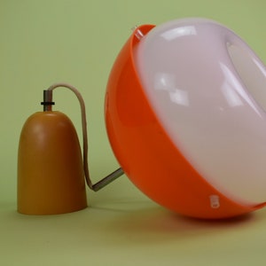 Könnte beinhalten: Eine moderne Pendelleuchte aus der Mitte des Jahrhunderts mit einem weißen und orangefarbenen Kunststoffschirm. Der Schirm ist eine Halbkugel mit einem Loch oben. Die Leuchte hat ein braunes Kabel und einen braunen Metallfuß.