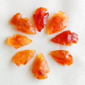May include: Eight orange and red arrowhead shaped stones arranged in a circle on a white surface.
