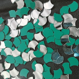 以下が含まれることがあります： 多数の扇形の装飾要素のクローズアップ。これらのパーツは、反射するシルバーとティールグリーンの混合で、暗い表面に散らばっています。形状は不規則で、テクスチャのあるモザイクのような外観を作り出しています。