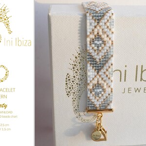 May include: A white and gray loom bracelet with a gold charm that says "Ini Ibiza" on a white box with the text "Ini Ibiza Jewelry" printed on it. The bracelet is 12.5 cm long and 1.5 cm wide.