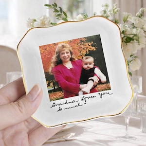 May include: A square white ceramic trinket dish with a gold-painted scalloped rim. The center features a color photograph of an adult and a baby outdoors in autumn, with the handwritten black text "Grandma Loves you so much." printed below.