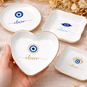 May include: A collection of white ceramic trinket dishes in various shapes: round, square, heart, and a wavy square. Each dish features a blue evil eye design and the name "oliver" in a script font, with gold trim.