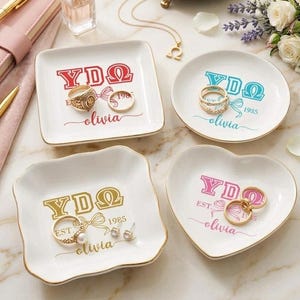 May include: Four white ceramic ring dishes in square, round, and heart shapes, each with gold trim. The dishes feature the Greek letters YΔΩ, the name "olivia", and the year "1985" in various colors. Rings and earrings are displayed on the dishes.