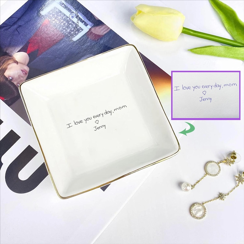 May include: A white square ceramic trinket dish with gold trim. The dish has the handwritten text "I love you every day, mom ♡ Jenny". A small card with the same text is also visible. A pair of earrings and a yellow tulip are in the background.