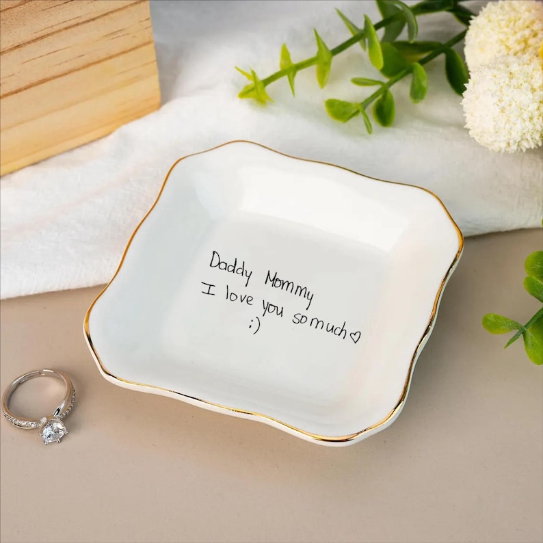 May include: A white ceramic ring dish with a gold trim and a handwritten message that reads "Daddy Mommy I love you so much :)". A silver ring with a large, clear stone sits next to the dish.