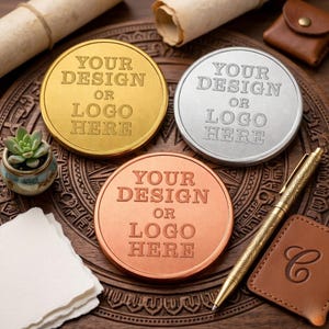 May include: Three metallic coins in gold, silver, and copper, each engraved with "YOUR DESIGN OR LOGO HERE." The coins rest on a carved wooden surface, accompanied by a pen, leather tag, and a small succulent plant in a pot. A scroll is in the background.