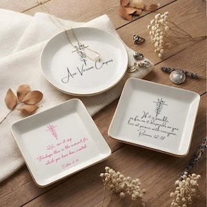 May include: Three white ceramic trinket dishes with gold-toned rims. One round dish features a floral cross and the text Ave Verum Corpus. Two square dishes feature floral crosses with the text Romans 12:18 and Esther 4:14.