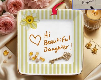 May include: A square ceramic trinket dish with a white center and light green striped border. The center features a hand-written brown script reading "Hi Beautiful Daughter!" with a heart icon. A gold-toned ornate key and small beads rest on the dish.
