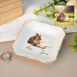 May include: White ceramic trinket dish with a gold rim, featuring a photo of two dogs. The names "Max" and "Juno" are written in script below the image. A diamond ring sits to the left of the dish.
