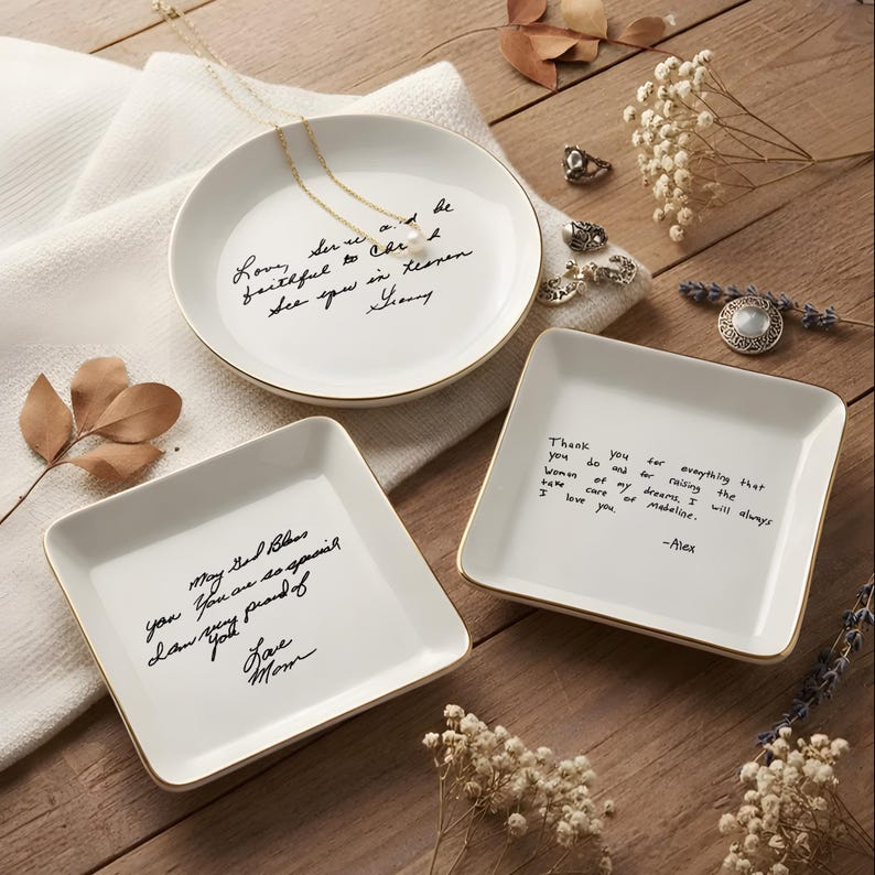 May include: Three white ceramic trinket dishes with gold rims and handwritten messages. One round dish and two square dishes are arranged on a wooden surface. A gold necklace and rings are also visible.