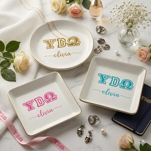 May include: Three white ceramic ring dishes with gold trim. Each dish has a different color of text: gold, pink, and teal. The text includes Greek letters and the name "olivia". A necklace, rings, and earrings are also present.