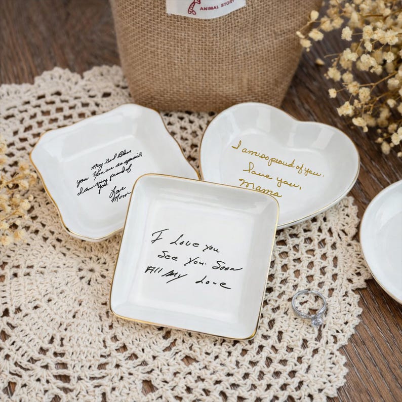 May include: White ceramic trinket dishes in various shapes, including square, heart, and irregular. Each dish has gold trim and handwritten messages in black or gold ink. A silver ring sits nearby.