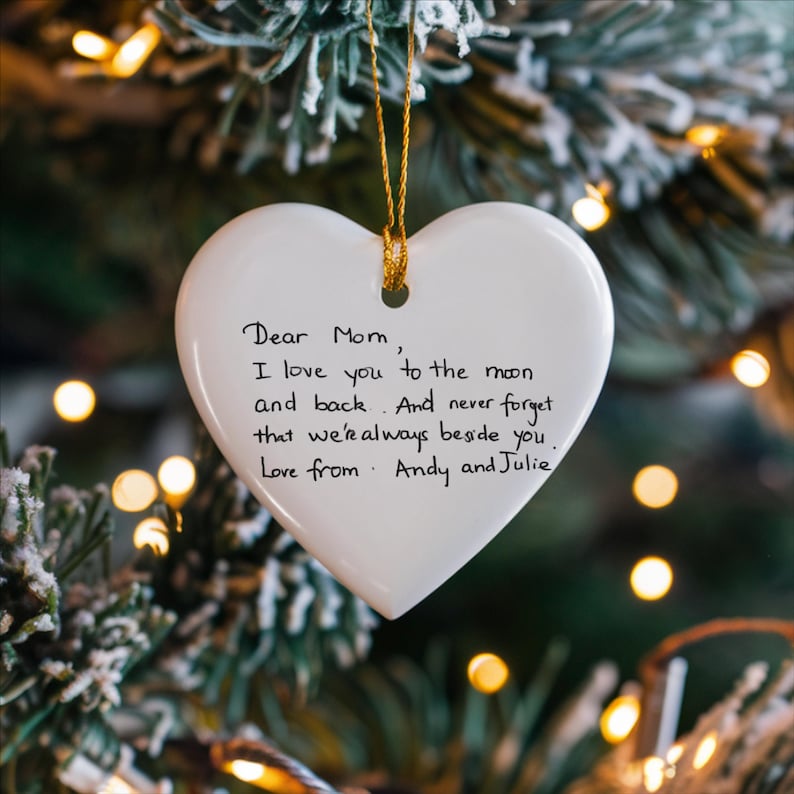 May include: White heart-shaped ornament with a gold string, hanging on a Christmas tree. The ornament has handwritten text that reads "Dear Mom, I love you to the moon and back. And never forget that we're always beside you. Love from Andy and Julie."