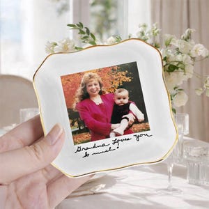 May include: A white ceramic dish with a gold rim, holding a photograph of a woman and a baby. The photo is inscribed with the words "Grandma Loves You so much!"