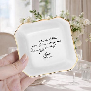 May include: A white ceramic trinket dish with a gold rim and a square shape. The dish has a handwritten message in black ink that reads "May God Bless you. You are so special. Love you. I am very proud of you. Love Mom."