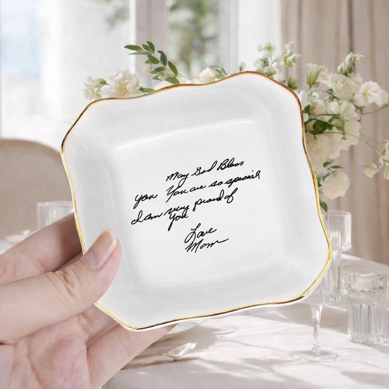 May include: A white ceramic trinket dish with a gold rim and a square shape. The dish has a handwritten message in black ink that reads "May God Bless you. You are so special. Love you. I am very proud of you. Love Mom."