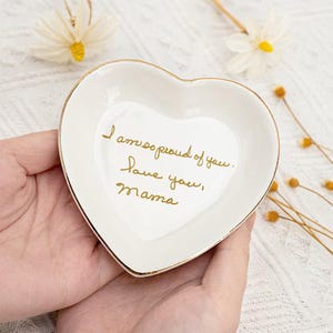 May include: A heart-shaped, white ceramic trinket dish with a gold rim. The dish has the handwritten text "I am so proud of you. Love you, mama" in gold. A decorative item for a desk or dresser.