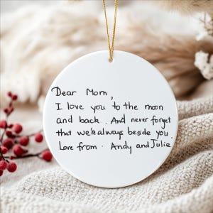 May include: A white, round ceramic ornament with a gold string for hanging. The ornament features handwritten text that reads "Dear Mom, I love you to the moon and back. And never forget that we're always beside you. Love from Andy and Julie."