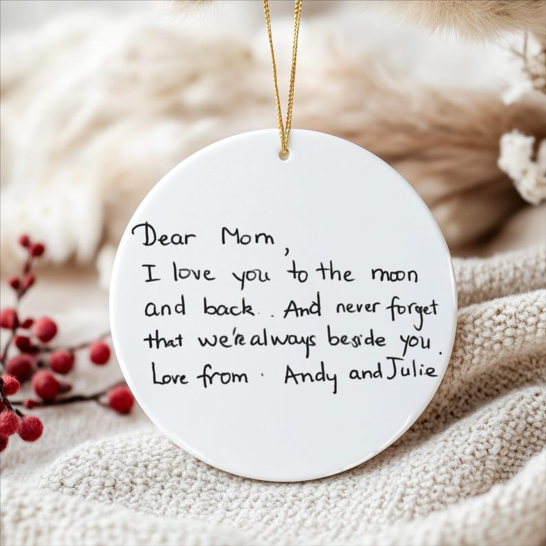 May include: A white, round ceramic ornament with a gold string for hanging. The ornament features handwritten text that reads "Dear Mom, I love you to the moon and back. And never forget that we're always beside you. Love from Andy and Julie."