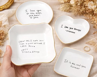 May include: Four white ceramic trinket dishes with gold rims. The dishes are in various shapes: round, square, heart, and rectangular. Each dish has handwritten text in black ink, including phrases like "I love you" and "forever."