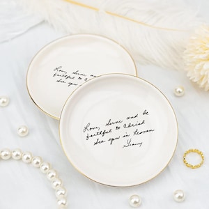 May include: Two white ceramic trinket dishes with gold rims. Each dish has black handwritten text. Pearls and a gold ring are scattered around the dishes on a white surface. The dishes are likely used for jewelry or small items.
