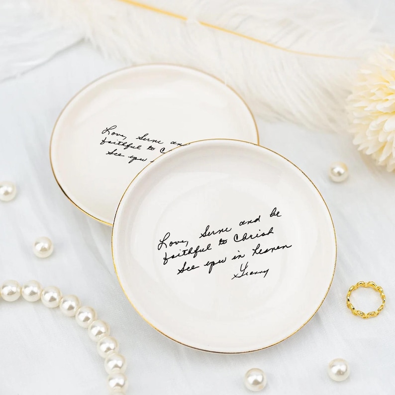 May include: Two white ceramic trinket dishes with gold rims. Each dish has black handwritten text. Pearls and a gold ring are scattered around the dishes on a white surface. The dishes are likely used for jewelry or small items.
