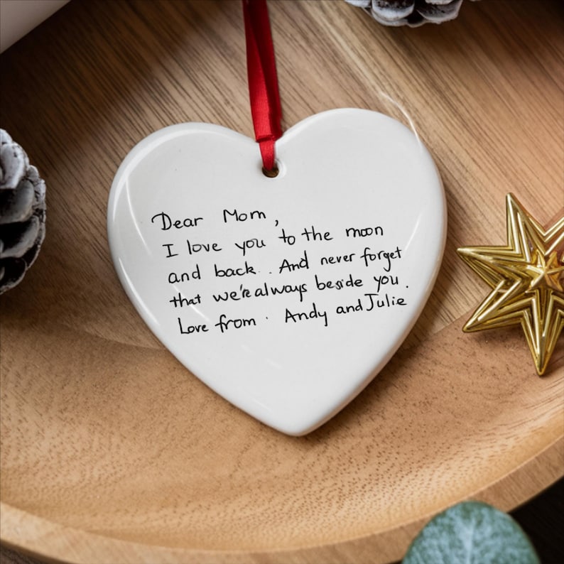 May include: A white heart-shaped ornament with a red ribbon for hanging. The ornament has handwritten text that reads, "Dear Mom, I love you to the moon and back. And never forget that we're always beside you. Love from Andy and Julie."