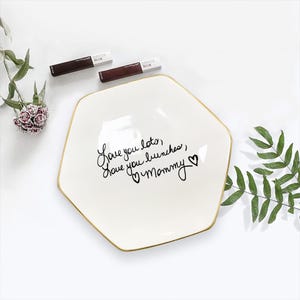 May include: A white, hexagonal ceramic dish with a gold rim, inscribed with the words "Love you lots, love you bunches, Mommy" in black script. Two tubes of Maybelline SuperStay Matte Ink lipstick are placed above the dish.