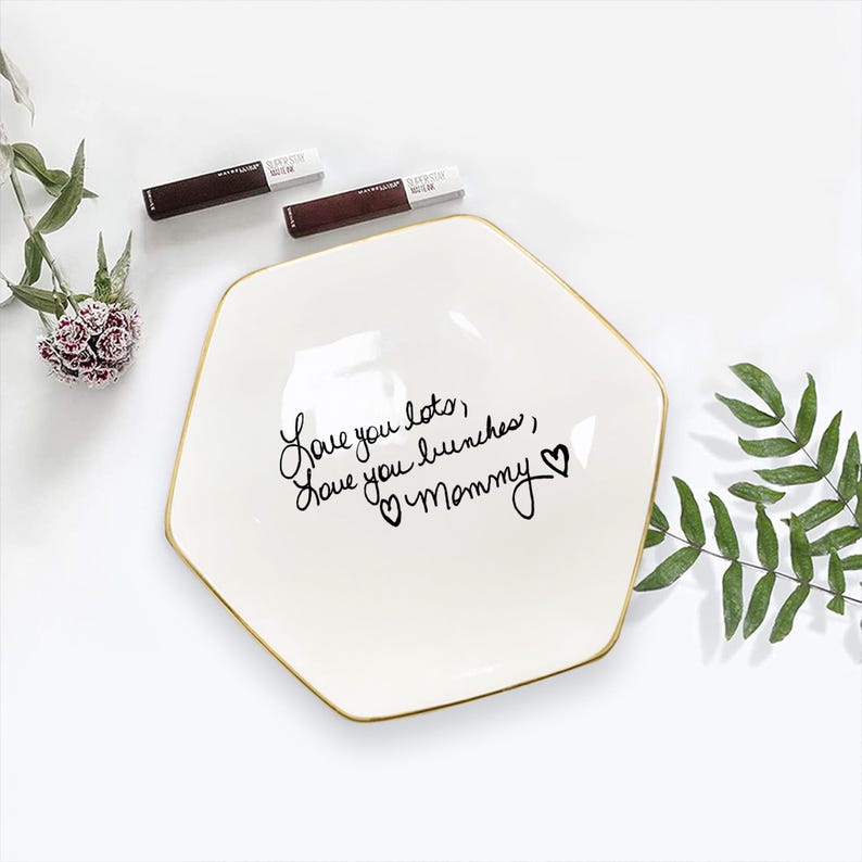 May include: A white, hexagonal ceramic dish with a gold rim, inscribed with the words "Love you lots, love you bunches, Mommy" in black script. Two tubes of Maybelline SuperStay Matte Ink lipstick are placed above the dish.