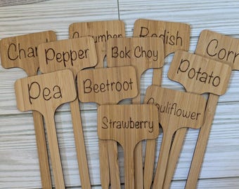 May include: A collection of bamboo garden markers with engraved names of vegetables and fruits, including "Char," "Pepper," "Pea," "Beetroot," "Strawberry," "Radish," "Corn," "Potato," and "Cauliflower."