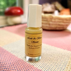 Puede incluir: Una peque&ntilde;a botella de vidrio de suero para pesta&ntilde;as y cejas con tapa blanca. La etiqueta dice "Herbs & Hearth" suero de Anna Morrow. La botella contiene un l&iacute;quido amarillo y est&aacute; sobre una superficie estampada rosa y blanca. Volumen neto: 10 ml.