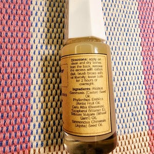 Puede incluir: Una peque&ntilde;a botella de vidrio transparente con tapa blanca, que contiene un l&iacute;quido marr&oacute;n claro. La etiqueta proporciona instrucciones y enumera ingredientes, incluyendo aceite de ricino y aceite de jojoba. La botella est&aacute; sobre una tela estampada.