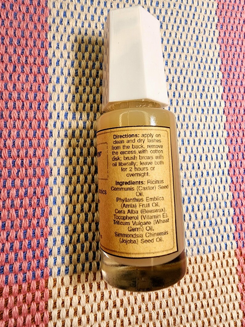Puede incluir: Una peque&ntilde;a botella de vidrio transparente con tapa blanca, que contiene un l&iacute;quido marr&oacute;n claro. La etiqueta proporciona instrucciones y enumera ingredientes, incluyendo aceite de ricino y aceite de jojoba. La botella est&aacute; sobre una tela estampada.