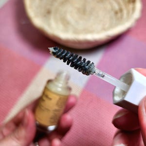 Puede incluir: Primer plano de un cepillo de r&iacute;mel con cerdas negras, sujeto contra un mango y una tapa blancos. Una peque&ntilde;a botella de l&iacute;quido se sostiene en la otra mano. Una cesta tejida se encuentra en el fondo sobre una superficie rosa y blanca.