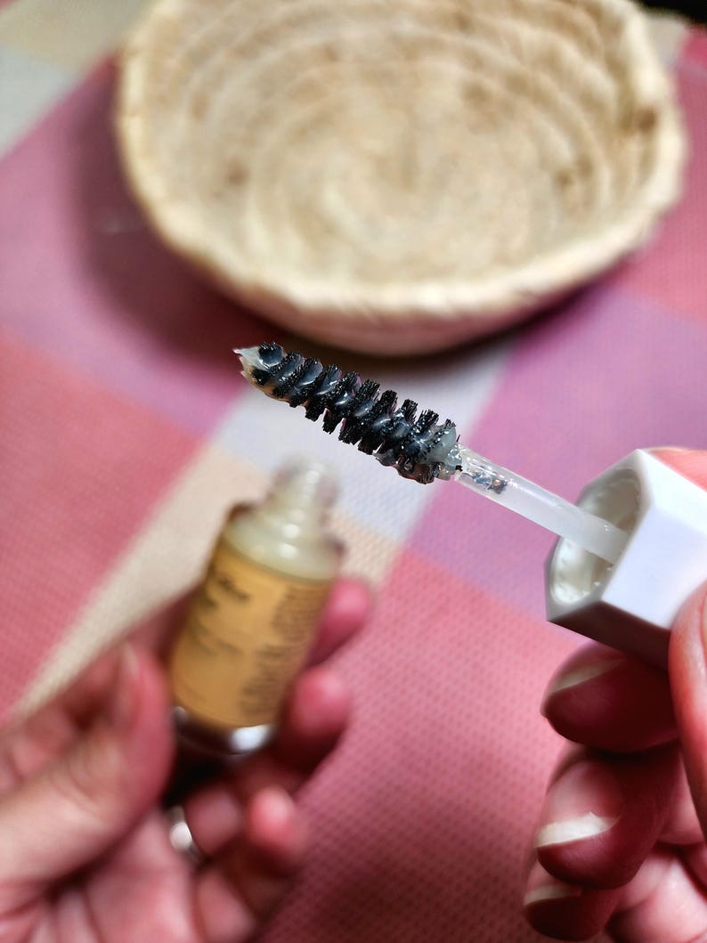 Puede incluir: Primer plano de un cepillo de r&iacute;mel con cerdas negras, sujeto contra un mango y una tapa blancos. Una peque&ntilde;a botella de l&iacute;quido se sostiene en la otra mano. Una cesta tejida se encuentra en el fondo sobre una superficie rosa y blanca.