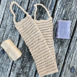 May include: A pair of natural fiber exfoliating bath scrubbers made of crocheted jute, with a bar of purple soap on a wooden surface.