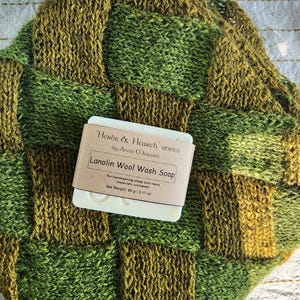May include: A handmade, green and brown knitted item with a checkerboard pattern. A bar of Lanolin Wool Wash Soap rests on the knitted item. The soap is labeled "Herbs & Hearth series by Anna Morwen" and has a net weight of 3.17 oz.