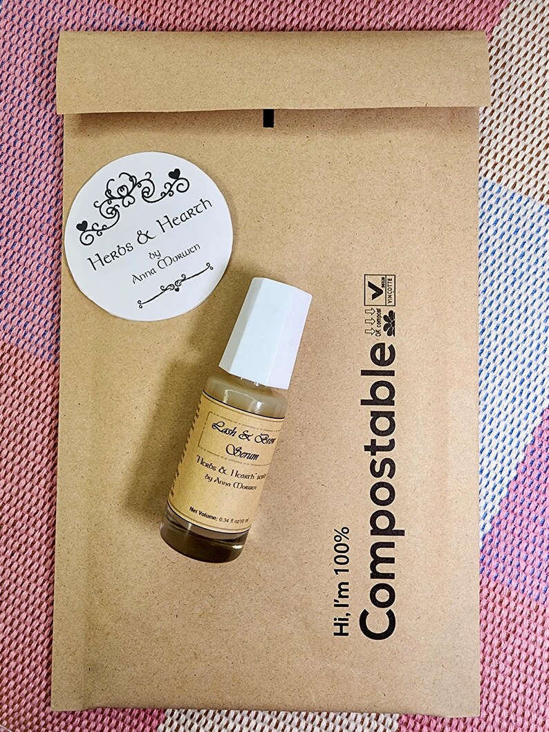 Puede incluir: Una peque&ntilde;a botella de vidrio de suero para pesta&ntilde;as y cejas con una tapa blanca se encuentra sobre un sobre de papel marr&oacute;n. El sobre tiene la palabra "Compostable" impresa. Tambi&eacute;n hay una pegatina redonda con las palabras "Herbs & Hearth".