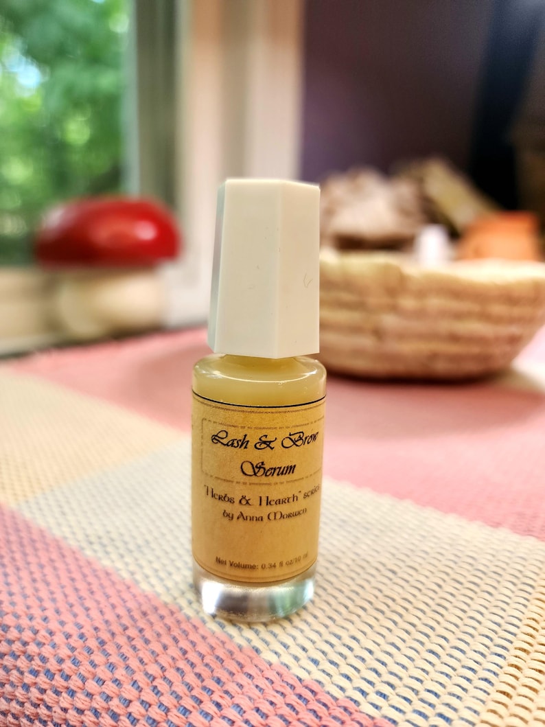 Puede incluir: Una peque&ntilde;a botella de vidrio de suero para pesta&ntilde;as y cejas con tapa blanca. La etiqueta dice "Herbs & Hearth" suero de Anna Morrow. La botella contiene un l&iacute;quido amarillo y est&aacute; sobre una superficie estampada rosa y blanca. Volumen neto: 10 ml.