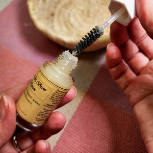 Puede incluir: Un frasco de vidrio transparente de s&eacute;rum para cejas con una tapa blanca y un aplicador de cepillo negro. El frasco tiene una etiqueta beige con texto. El cepillo se est&aacute; sacando del frasco. Una cesta tejida est&aacute; en el fondo.