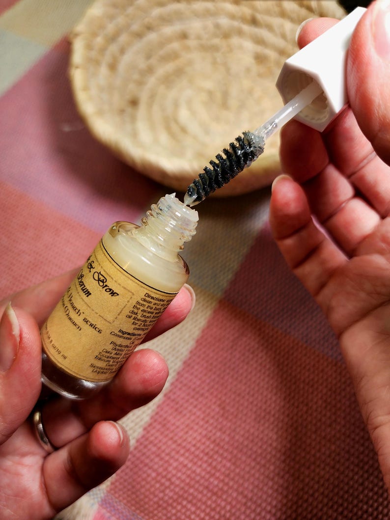 Puede incluir: Un frasco de vidrio transparente de s&eacute;rum para cejas con una tapa blanca y un aplicador de cepillo negro. El frasco tiene una etiqueta beige con texto. El cepillo se est&aacute; sacando del frasco. Una cesta tejida est&aacute; en el fondo.