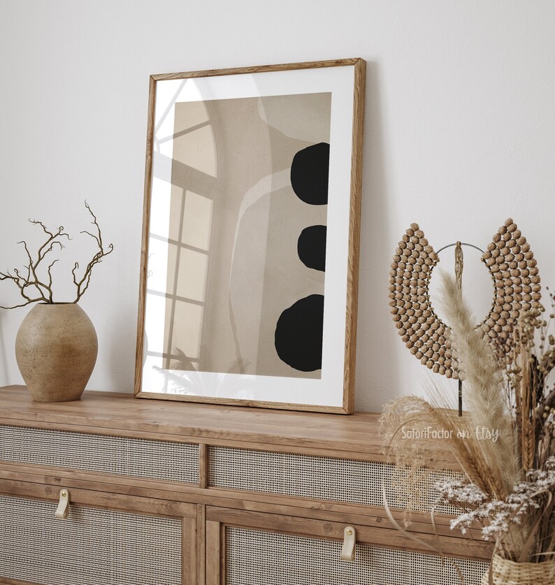 May include: A framed abstract art print with a beige background and three black circles. The print is hanging on a white wall above a wooden cabinet with woven rattan doors.